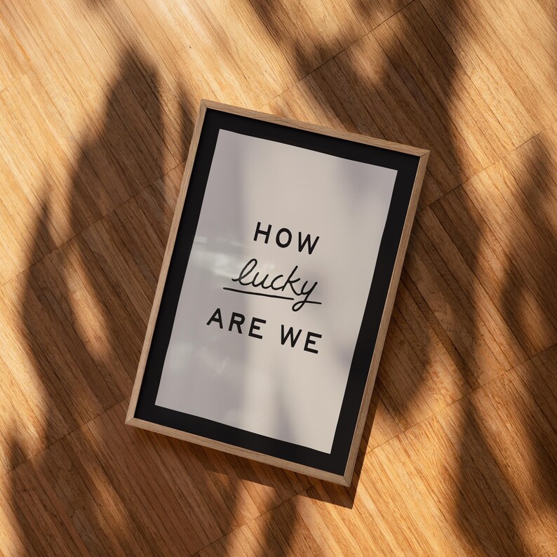Puede incluir: Una impresi&oacute;n enmarcada con las palabras "HOW lucky ARE WE" en negro y fuente de escritura. La impresi&oacute;n tiene un borde negro y un fondo de color claro. El marco es de color madera clara, y la impresi&oacute;n est&aacute; sobre una superficie de madera.