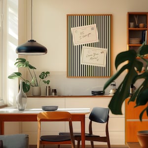 May include: A modern interior scene featuring a dining table with two chairs, a decorative framed board with postcards, and a hanging black pendant light. The board has a green and white striped pattern. The postcards read "PS I love you" and "Love You More!"
