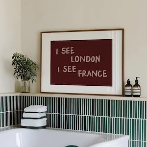 May include: A rectangular wall art print in a thin wooden frame featuring a dark maroon background with the light pink text I SEE LONDON I SEE FRANCE displayed in four lines. The artwork is positioned above a white bathtub with green tiles.