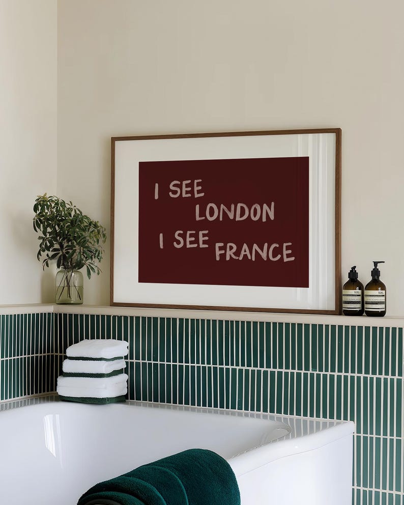 May include: A rectangular wall art print in a thin wooden frame featuring a dark maroon background with the light pink text I SEE LONDON I SEE FRANCE displayed in four lines. The artwork is positioned above a white bathtub with green tiles.