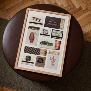 May include: A framed art print featuring various matchbox designs. The designs include text like "777", "The End", and "Lucky You", along with images of a car and a horseshoe. The frame is light-colored, and the artwork is displayed on a white background.