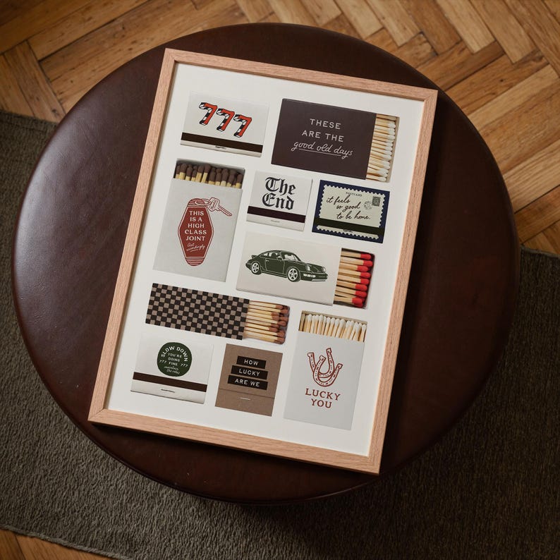 May include: A framed art print featuring various matchbox designs. The designs include text like "777", "The End", and "Lucky You", along with images of a car and a horseshoe. The frame is light-colored, and the artwork is displayed on a white background.