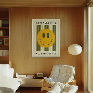 May include: A framed poster with a yellow smiley face on a green striped background. The text "ACTUALLY IT IS" and "ALL FUN + GAMES" is printed above and below the smiley face. The poster is hanging on a light wood paneled wall.