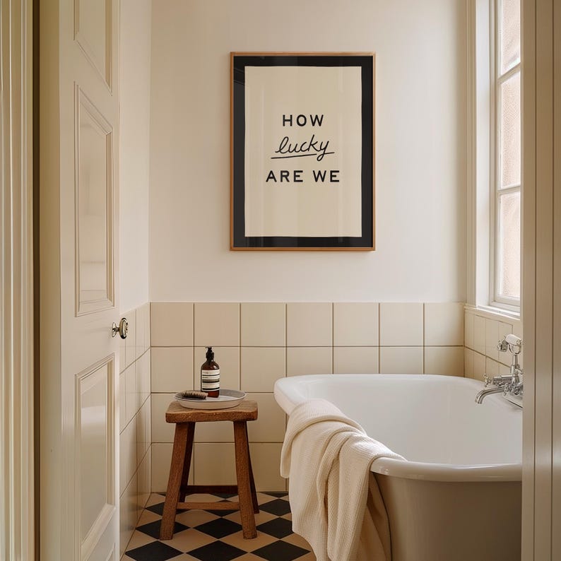 Puede incluir: Interior de ba&ntilde;o con un cuadro enmarcado con el texto "HOW lucky ARE WE". La habitaci&oacute;n incluye una ba&ntilde;era blanca, un taburete de madera y un frasco de jab&oacute;n. El suelo tiene un patr&oacute;n de cuadros blancos y negros.