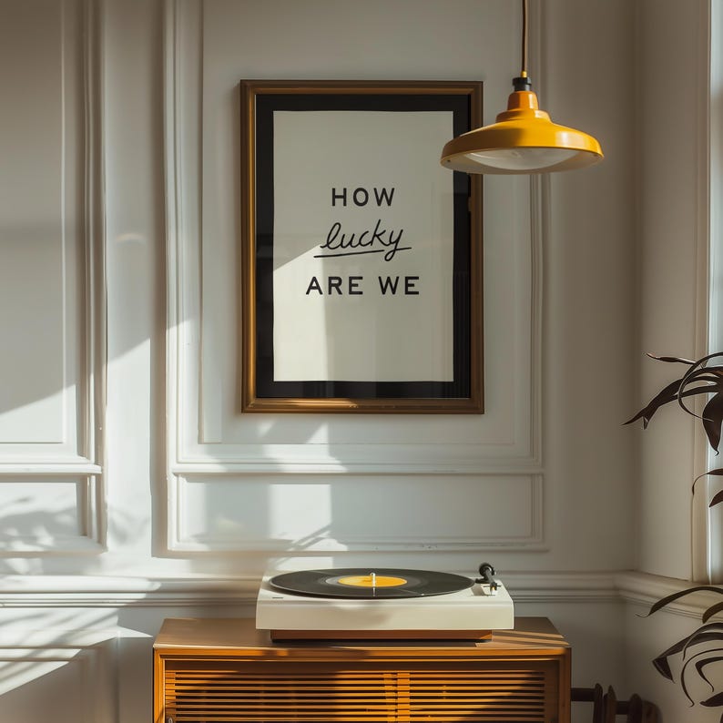 Puede incluir: Una impresi&oacute;n enmarcada con el texto "HOW lucky ARE WE" cuelga sobre un tocadiscos en un mueble de madera. La impresi&oacute;n tiene un marco dorado y un passe-partout negro. Una l&aacute;mpara colgante amarilla cuelga del techo.