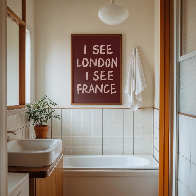 May include: A bathroom scene featuring a framed burgundy sign with the text "I SEE LONDON I SEE FRANCE". The room has white tiled walls, a bathtub, a sink, and a potted plant. A white towel hangs on the wall.