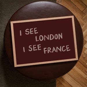 May include: A rectangular wall art piece in a light wood frame featuring a deep maroon background with the white text I SEE LONDON I SEE FRANCE written in a casual, hand-lettered style across two lines, centered within the frame.