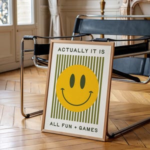 May include: A framed art print with a yellow smiley face and the text "ACTUALLY IT IS" above and "ALL FUN + GAMES" below. The background features vertical green stripes. The frame is a light brown color.