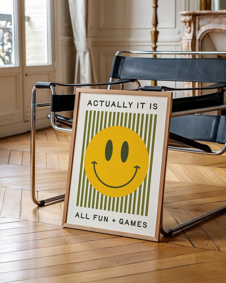 May include: A framed art print with a yellow smiley face and the text "ACTUALLY IT IS" above and "ALL FUN + GAMES" below. The background features vertical green stripes. The frame is a light brown color.