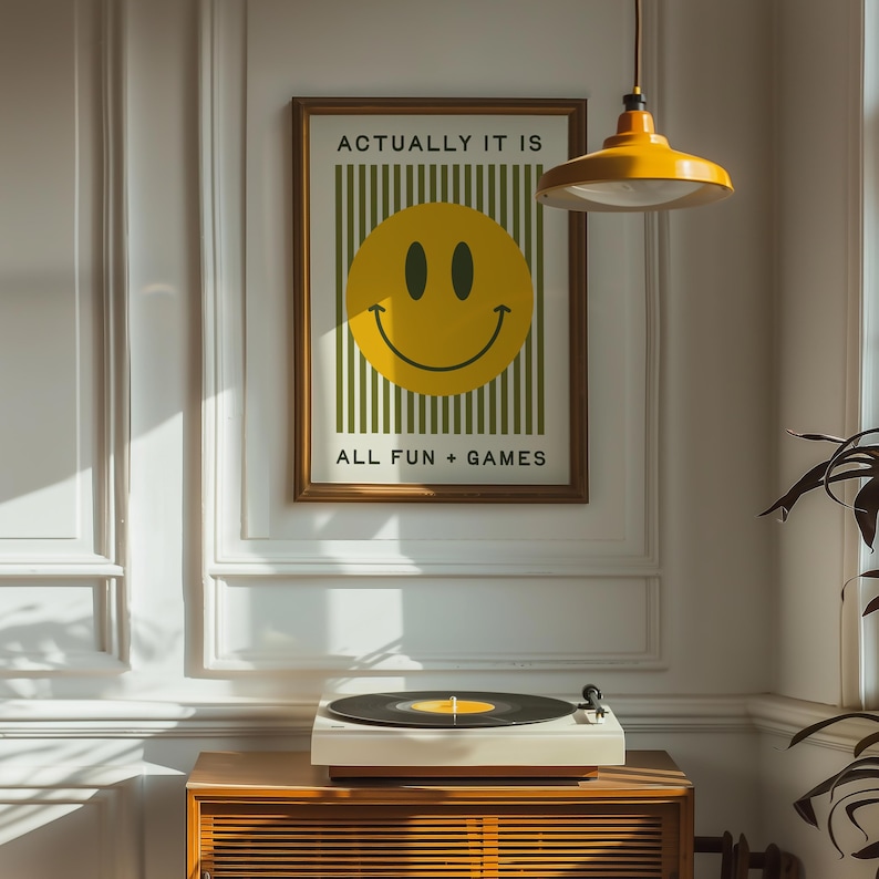 May include: A framed poster with the text "ACTUALLY IT IS ALL FUN + GAMES" and a large yellow smiley face. A white record player sits on a wooden cabinet. A yellow pendant light hangs above.