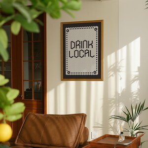May include: A framed wall art piece with the words "DRINK LOCAL" in a black and white mosaic tile design. The frame is a warm brown color. The art hangs on a cream-colored wall above a brown leather couch.
