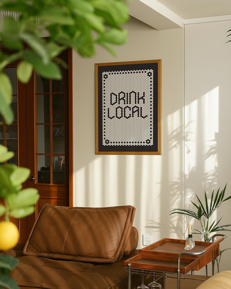May include: A framed wall art piece with the words "DRINK LOCAL" in a black and white mosaic tile design. The frame is a warm brown color. The art hangs on a cream-colored wall above a brown leather couch.
