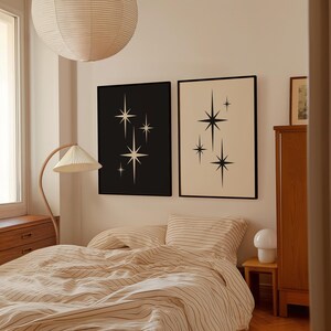May include: Two framed art prints with a mid-century modern design. One print has a black background with white starburst shapes, and the other has a cream background with black starburst shapes. The prints are displayed above a bed with striped bedding.