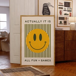 May include: A framed poster with a yellow smiley face and the text "ACTUALLY IT IS ALL FUN + GAMES." The background has green vertical stripes. Other framed art pieces are visible in the background.