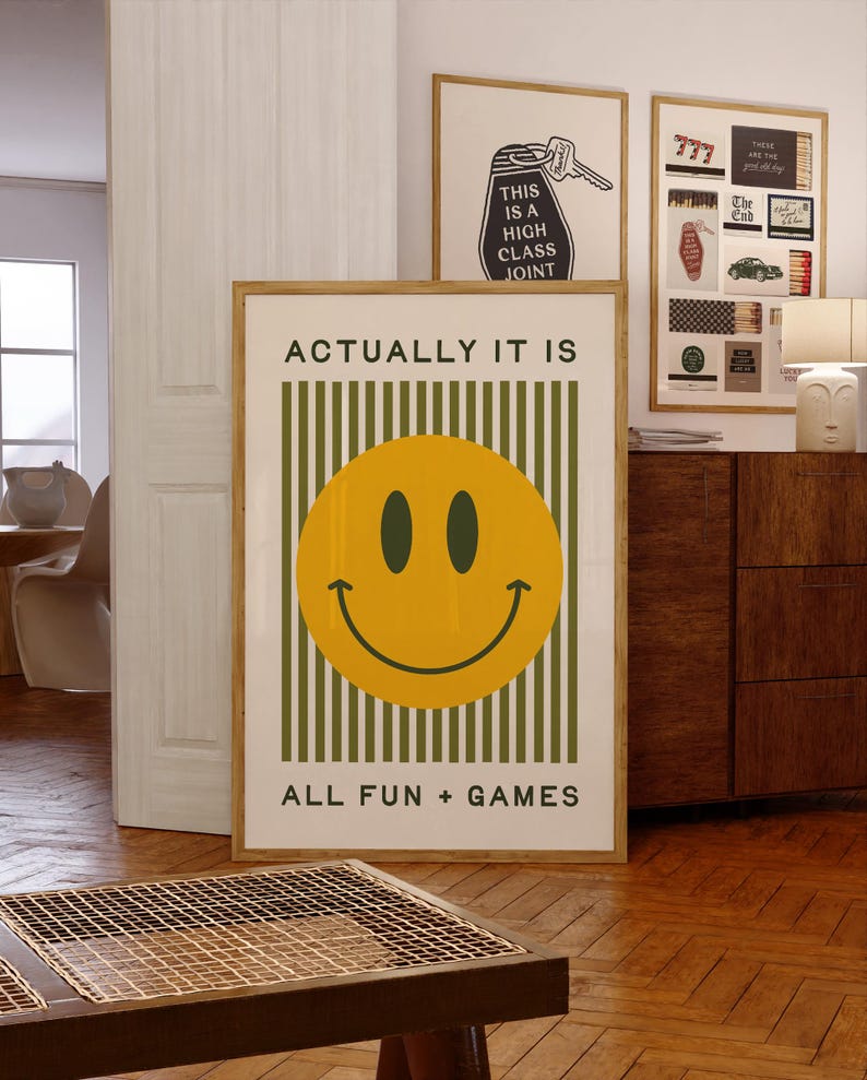 May include: A framed poster with a yellow smiley face and the text "ACTUALLY IT IS ALL FUN + GAMES." The background has green vertical stripes. Other framed art pieces are visible in the background.