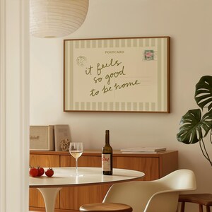 May include: A framed postcard print with the text "it feels so good to be home" in a dining room setting. The room includes a round table, wine bottle, wine glass, and tomatoes.