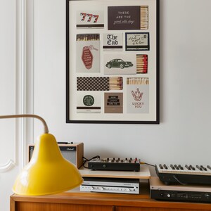 May include: A framed art piece featuring various matchbox designs with text like "777", "The End", and "Lucky You". The artwork is displayed above a wooden cabinet with a yellow lamp, audio equipment, and a keyboard.