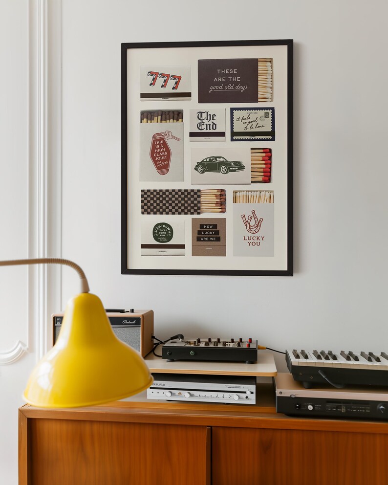 May include: A framed art piece featuring various matchbox designs with text like "777", "The End", and "Lucky You". The artwork is displayed above a wooden cabinet with a yellow lamp, audio equipment, and a keyboard.