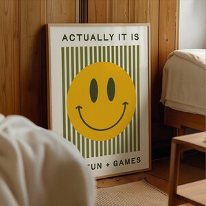 May include: A framed poster with a yellow smiley face and the text "ACTUALLY IT IS" above and "FUN + GAMES" below. The background has vertical green stripes. The frame is a light brown color.