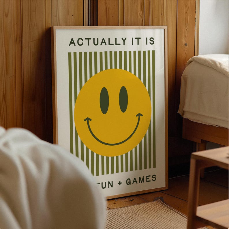 May include: A framed poster with a yellow smiley face and the text "ACTUALLY IT IS" above and "FUN + GAMES" below. The background has vertical green stripes. The frame is a light brown color.