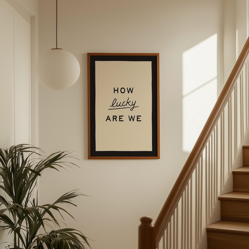 Puede incluir: Un cuadro enmarcado con las palabras "HOW lucky ARE WE" en escritura negra. La impresi&oacute;n tiene un fondo crema, un borde negro y un marco marr&oacute;n. La obra de arte cuelga en una pared blanca cerca de una escalera.