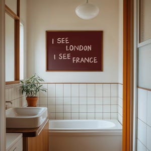 May include: A rectangular wall art piece in a wooden frame featuring a dark maroon background with the white text I SEE LONDON I SEE FRANCE displayed in a casual, hand-lettered style, mounted on a white wall above a white bathtub and sink.