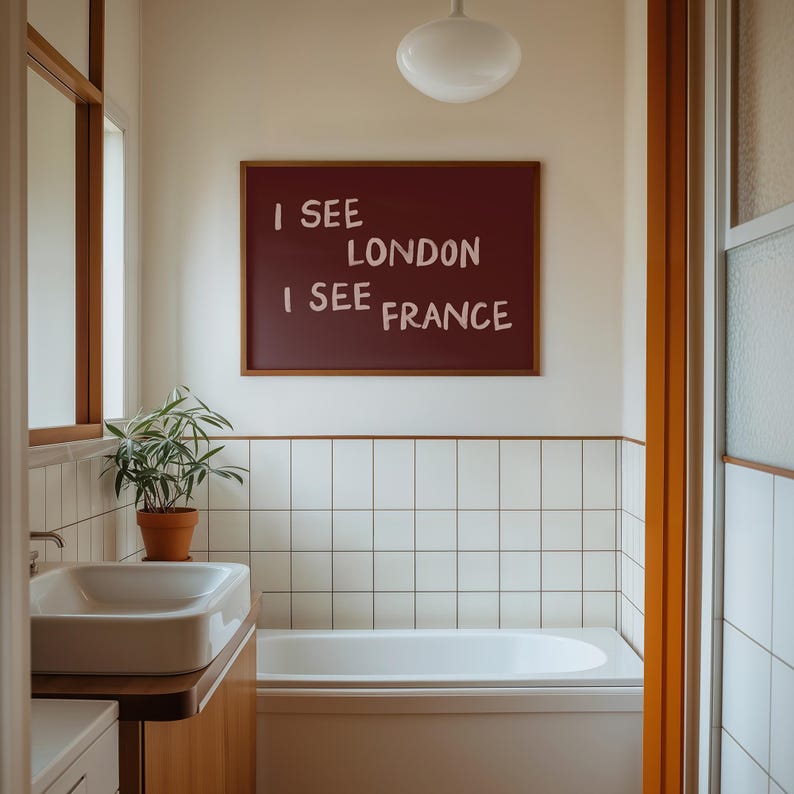May include: A rectangular wall art piece in a wooden frame featuring a dark maroon background with the white text I SEE LONDON I SEE FRANCE displayed in a casual, hand-lettered style, mounted on a white wall above a white bathtub and sink.