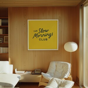 以下が含まれることがあります： 明るい木製パネルと、黄色の背景に黒い筆記体で「The Slow Mornings Club」と書かれた白いフレームのポスターがあるモダンなリビングルーム。白い椅子とソファが見えます。