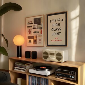 May include: Two framed wall art prints with light wood frames. One print features a collage of matchbook designs, and the other displays the black text THIS IS A HIGH CLASS JOINT and red text ACT ACCORDINGLY on a cream background.
