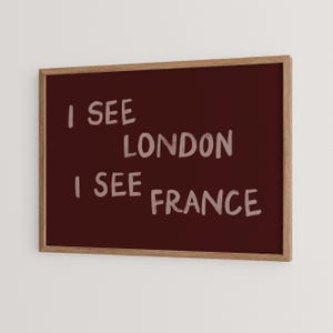 May include: A rectangular wall art piece in a light wood frame featuring a deep burgundy background. The text I SEE LONDON I SEE FRANCE is printed in a light pink, hand-lettered style font across the center of the dark red surface.