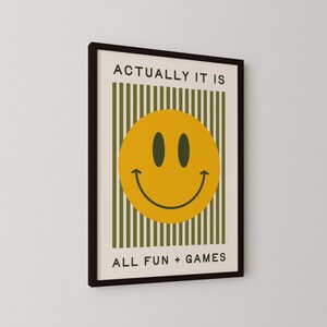 May include: Framed wall art featuring a yellow smiley face with black eyes and a curved mouth. The artwork includes the text "ACTUALLY IT IS" above the smiley face and "ALL FUN + GAMES" below. The background has vertical green stripes.