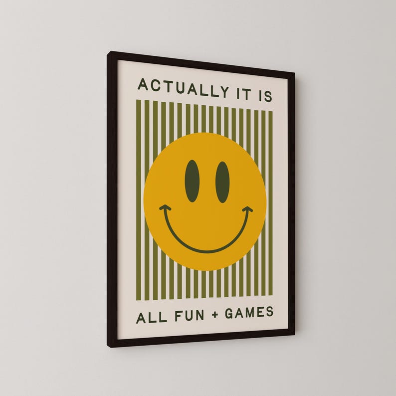May include: Framed wall art featuring a yellow smiley face with black eyes and a curved mouth. The artwork includes the text "ACTUALLY IT IS" above the smiley face and "ALL FUN + GAMES" below. The background has vertical green stripes.