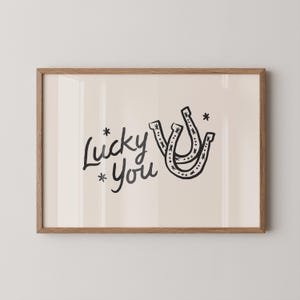Może przedstawiać: Oprawiony wydruk artystyczny z napisem "Lucky You" w czarnym skrypcie, kt&oacute;remu towarzyszą dwie czarne ilustracje podk&oacute;w. Grafika ma jasnobeżowe tło i jest oprawiona w ramę z naturalnego drewna.
