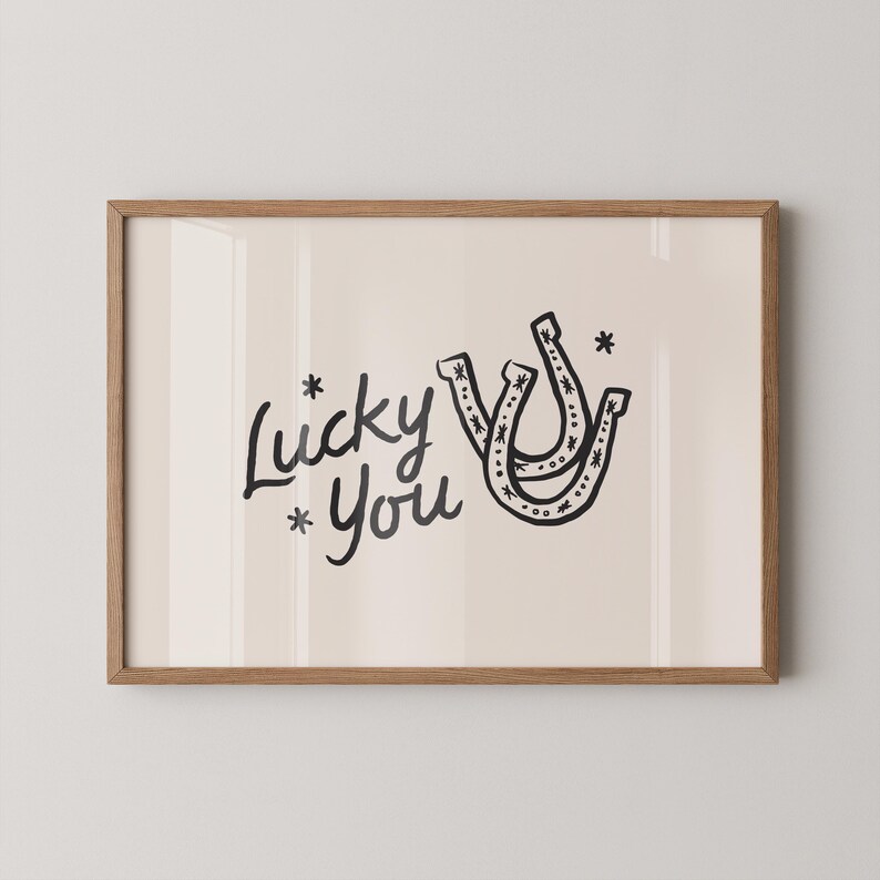 Może przedstawiać: Oprawiony wydruk artystyczny z napisem "Lucky You" w czarnym skrypcie, kt&oacute;remu towarzyszą dwie czarne ilustracje podk&oacute;w. Grafika ma jasnobeżowe tło i jest oprawiona w ramę z naturalnego drewna.