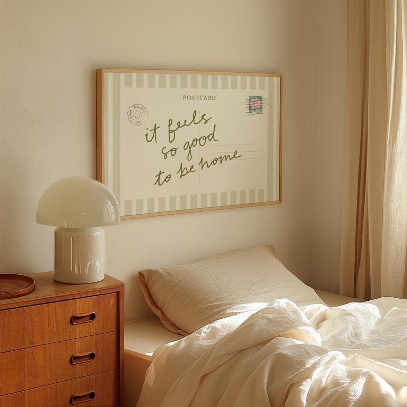 May include: A framed postcard print with the text "it feels so good to be home" hangs above a bed. A white mushroom-shaped lamp sits on a wooden nightstand. The bed has a cream-colored comforter and pillow.