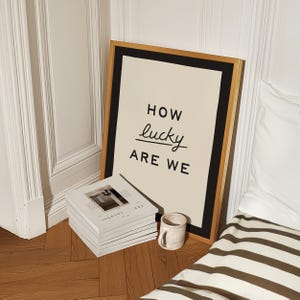 Puede incluir: Un cuadro enmarcado con el texto "HOW lucky ARE WE" en letras negras. El cuadro tiene un fondo color crema y un marco negro y dorado. Una pila de libros blancos y una peque&ntilde;a taza de cer&aacute;mica est&aacute;n frente al cuadro.