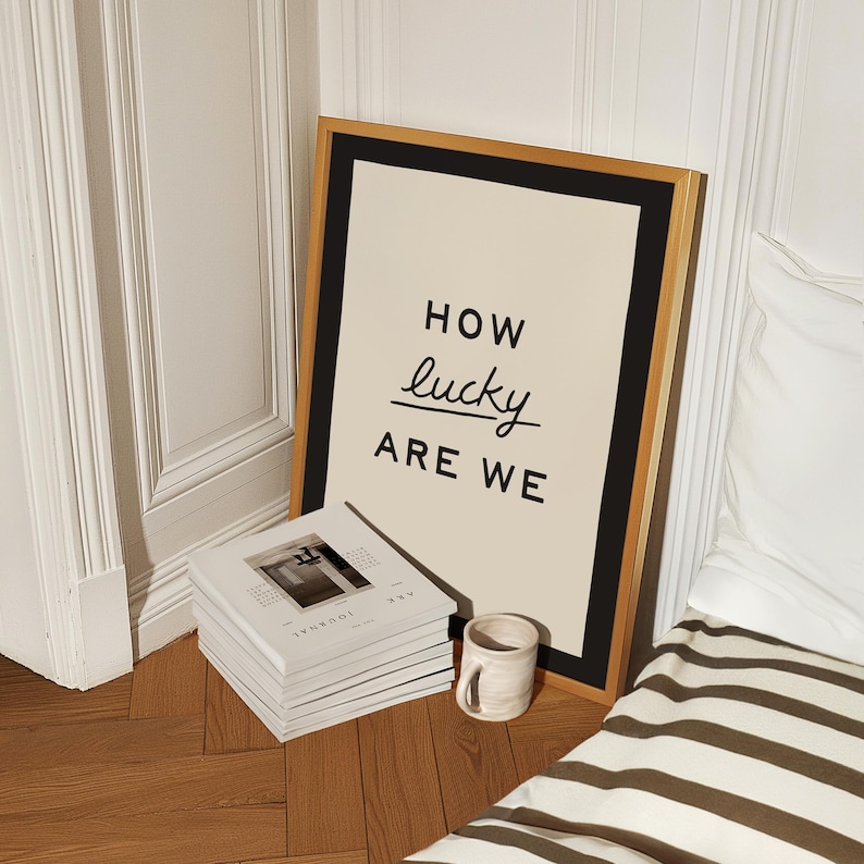 Puede incluir: Un cuadro enmarcado con el texto "HOW lucky ARE WE" en letras negras. El cuadro tiene un fondo color crema y un marco negro y dorado. Una pila de libros blancos y una peque&ntilde;a taza de cer&aacute;mica est&aacute;n frente al cuadro.