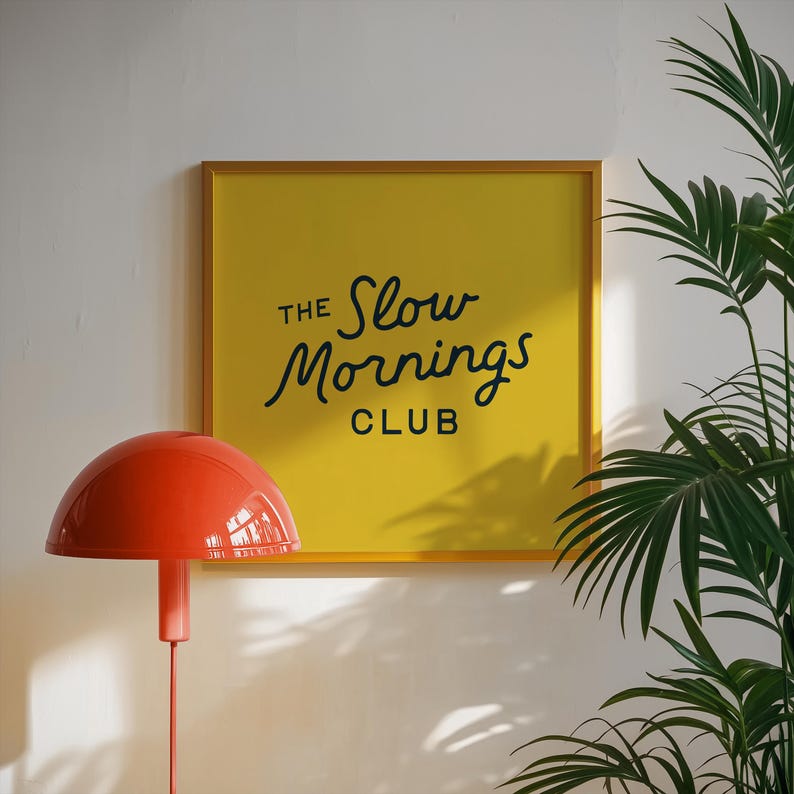 以下が含まれることがあります： 「The Slow Mornings Club」の黒い文字が書かれた、額縁に入った黄色の正方形のプリント。オレンジ色のキノコ型のランプが前に立っています。背景には緑の葉の植物があります。