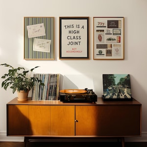 May include: A wooden sideboard holds a record player with an orange vinyl, a stack of records, and a potted plant. Above, three framed prints hang on the wall: one with green stripes and notes, one with the text THIS IS A HIGH CLASS JOINT ACT ACCORDINGLY, and one with various small graphic icons.
