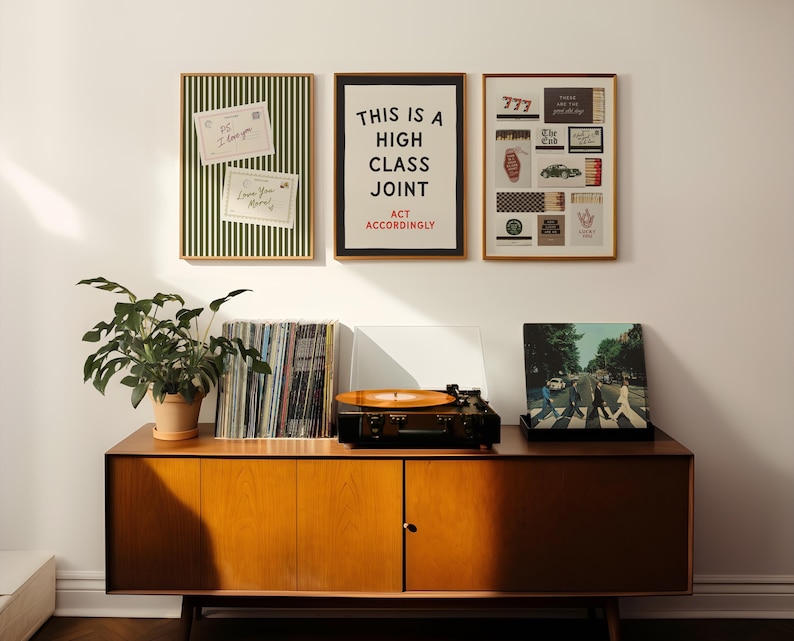 May include: A wooden sideboard holds a record player with an orange vinyl, a stack of records, and a potted plant. Above, three framed prints hang on the wall: one with green stripes and notes, one with the text THIS IS A HIGH CLASS JOINT ACT ACCORDINGLY, and one with various small graphic icons.