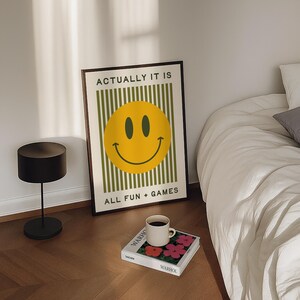 May include: A framed poster with a yellow smiley face on a green striped background. The text "ACTUALLY IT IS" and "ALL FUN + GAMES" is displayed. A black lamp, a coffee cup, and a book are also visible.