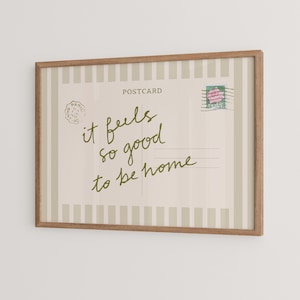 May include: A framed postcard print with a light beige background and vertical stripes. The text "it feels so good to be home" is written in green script. A postage stamp with a pink rose is in the upper right corner.