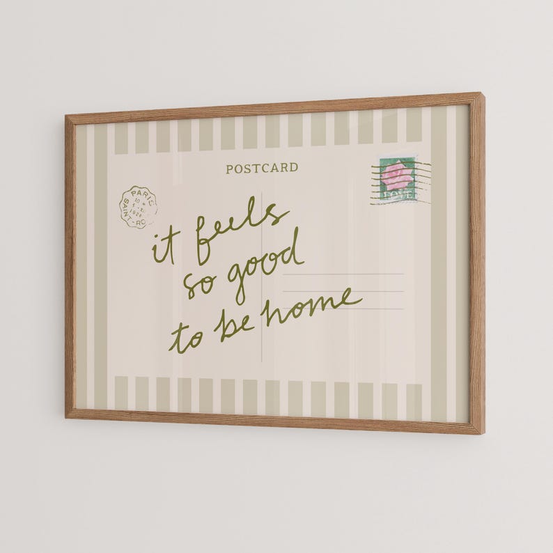 May include: A framed postcard print with a light beige background and vertical stripes. The text "it feels so good to be home" is written in green script. A postage stamp with a pink rose is in the upper right corner.