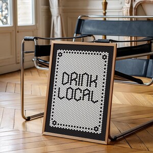 May include: A framed sign with the words "DRINK LOCAL" in bold, black letters. The sign has a black and white mosaic tile design and a wooden frame. The sign is leaning against a chair in a room with hardwood floors.