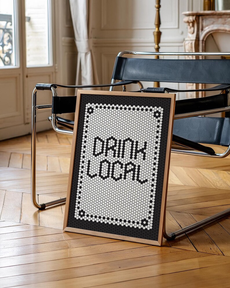 May include: A framed sign with the words "DRINK LOCAL" in bold, black letters. The sign has a black and white mosaic tile design and a wooden frame. The sign is leaning against a chair in a room with hardwood floors.