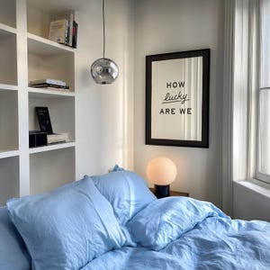 Puede incluir: Interior de dormitorio con ropa de cama y almohadas azul claro. Una impresi&oacute;n enmarcada en la pared dice "HOW lucky ARE WE". Una l&aacute;mpara esf&eacute;rica se encuentra en una mesita de noche. Una l&aacute;mpara colgante cromada cuelga del techo. Se ven estanter&iacute;as.