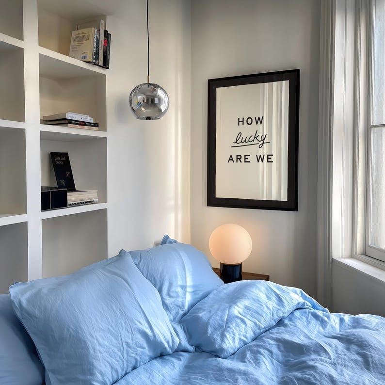 Puede incluir: Interior de dormitorio con ropa de cama y almohadas azul claro. Una impresi&oacute;n enmarcada en la pared dice "HOW lucky ARE WE". Una l&aacute;mpara esf&eacute;rica se encuentra en una mesita de noche. Una l&aacute;mpara colgante cromada cuelga del techo. Se ven estanter&iacute;as.