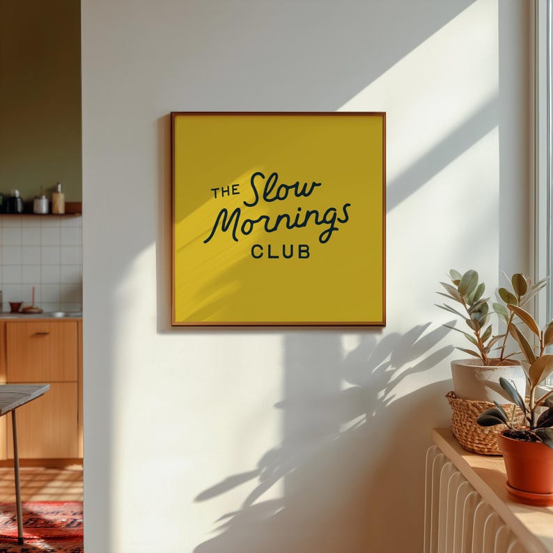 以下が含まれることがあります： 黄色い背景に黒い文字で「The Slow Mornings Club」と書かれた額入りの壁掛けアート。フレームは茶色です。白い壁に掛けられており、近くの窓台には鉢植えの植物があります。
