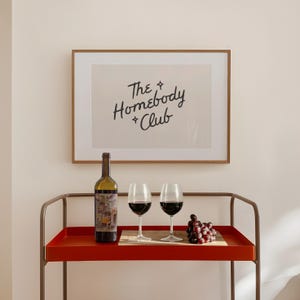 May include: A framed print with the text "The Homebody Club" hangs above a red serving cart. The cart holds a wine bottle, two wine glasses filled with red wine, and a bunch of grapes. The cart has a brown metal frame.