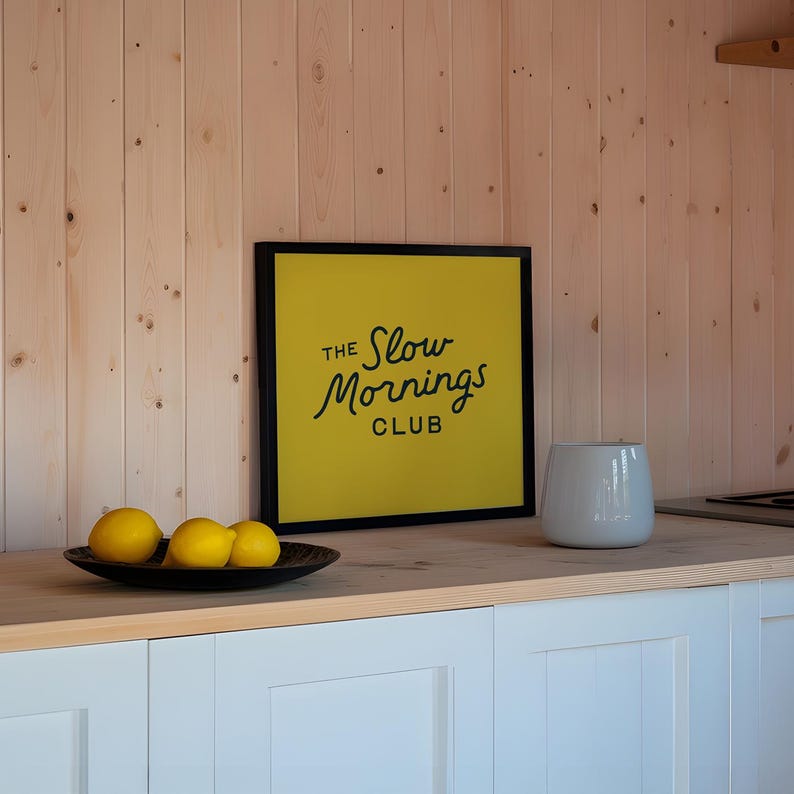 以下が含まれることがあります： 「The Slow Mornings Club」と書かれた黒いフレームの黄色い看板があるキッチン。灰色のセラミックの花瓶と、3つのレモンが入った濃い木製のボウルがカウンターに置かれています。キャビネットは白です。